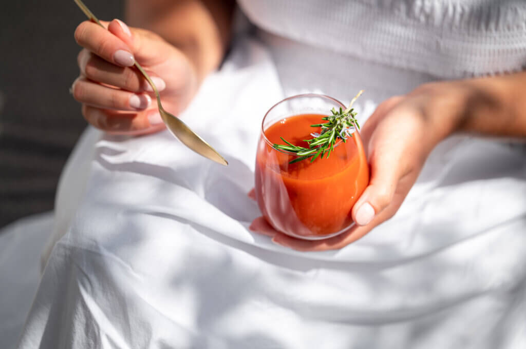 Frau fastet im Sommer mit der Fastenbox und hält ein Glas gekühlte Tomatensuppe mit einem Rosmarin-Zweig in der Hand